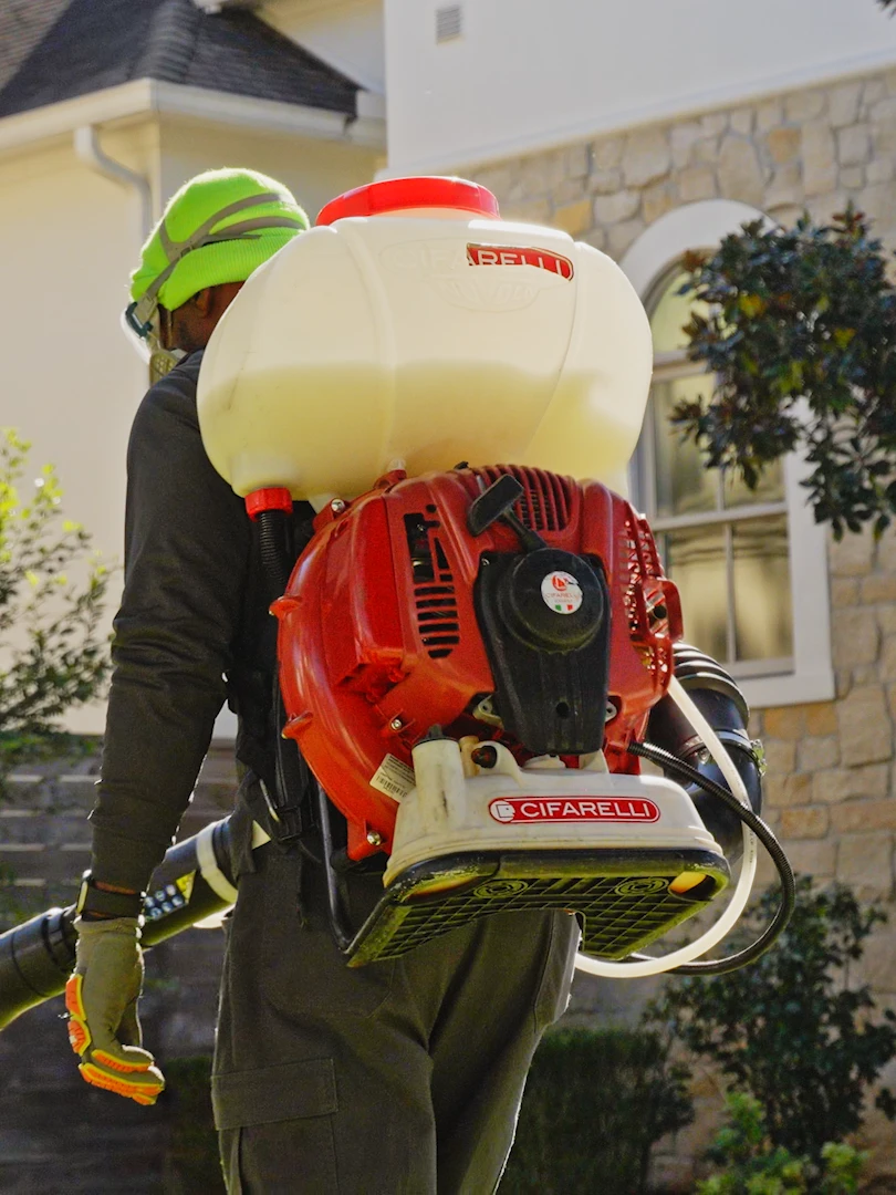 Mosquito control technician wearing a backpack mist blower in the yard of a stone-sided home, ready to apply a barrier treatment around the house and landscaping.